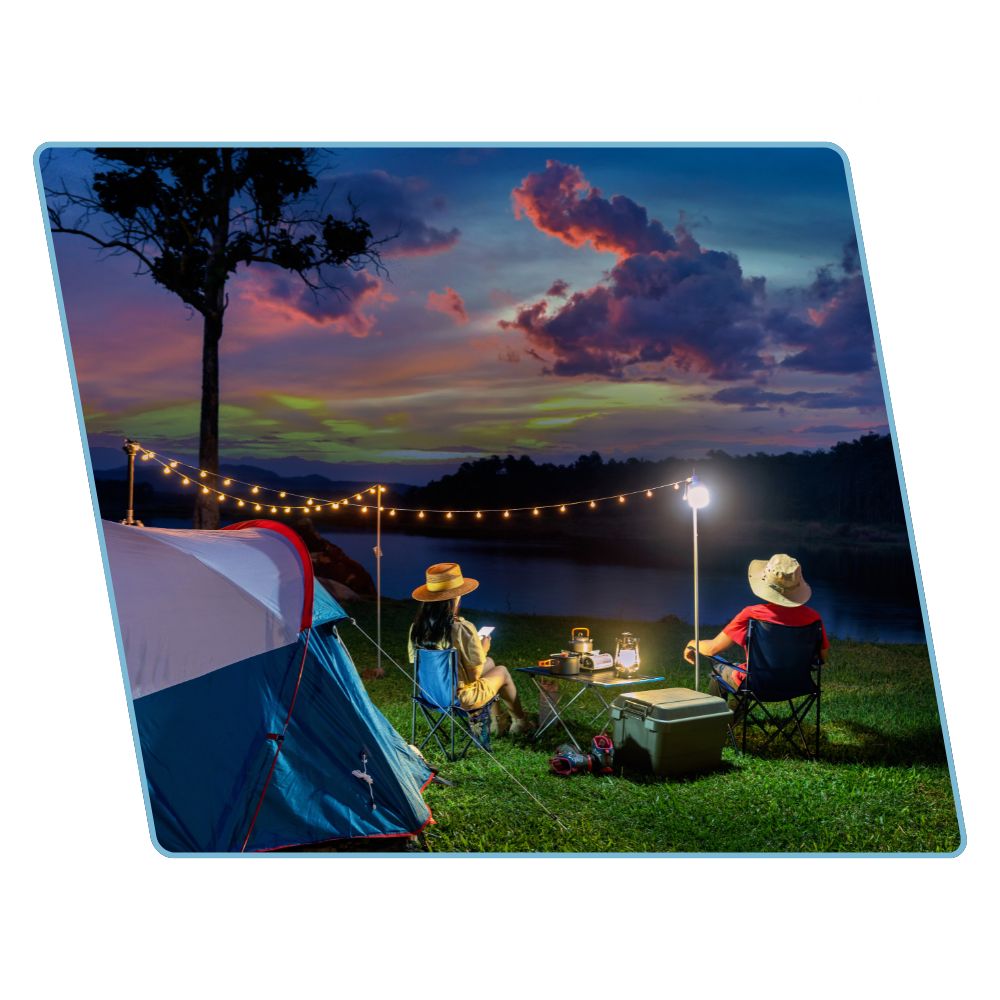 A square product shot of a printed mat or cushion featuring a camping scene at dusk, with a blue and white tent, two people sitting in chairs under string lights and a glowing lamp, a small table with bottles and a cooler, and a dramatic sunset sky over a lake. The item is shown on a plain white background with a slight drop shadow.